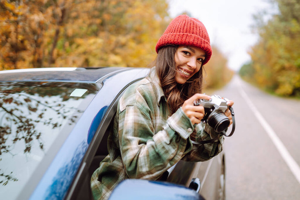 Beautiful woman takes pictures on the camerafrom the car window. Smiling woman enjoying autumn weather. Rest, relaxation, lifestyle concept. - 写真・画像