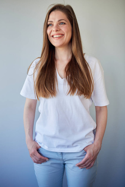 Smiling woman in white shirt looking away and keeps hands in pockets. Happy girl with long brown hair, isolated portrait. - Фото, зображення