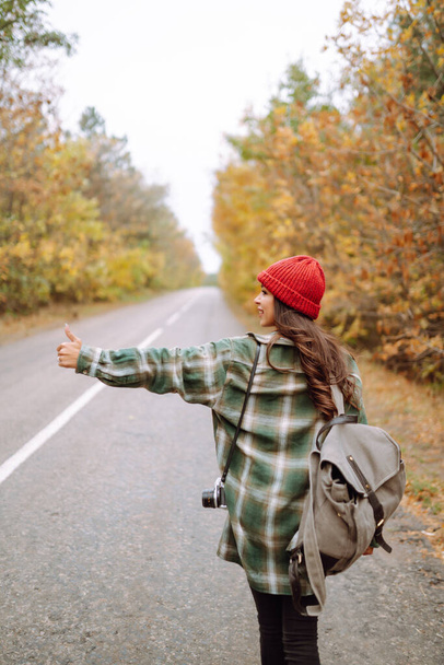 Young female hitchhiker by the roadside among autumn forest during fall season. Travel woman hitchhiking. Rest, relaxation, travel, lifestyle concept. - 写真・画像