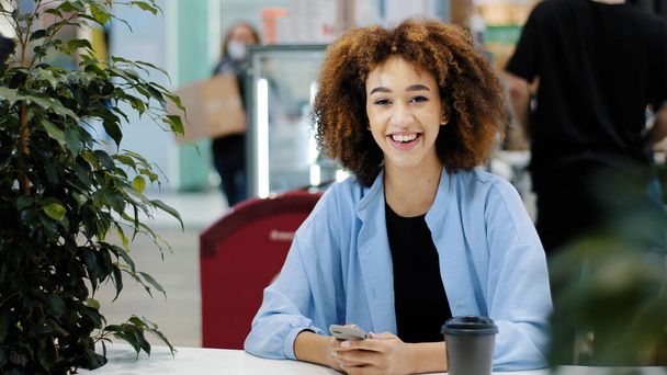 Young beautiful girl teenager millennial African American woman with curly hair sits in cafe looking at mobile phone rejoices wins gets victory good result winning in online game luck success triumph - Photo, Image