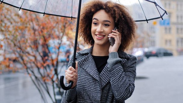 Ευτυχισμένο ενεργό φιλικό σγουρά μαλλιά κορίτσι της Αφρικής αμερικανική γυναίκα με διαφανή ομπρέλα στην πόλη σε θυελλώδη βροχερό καιρό απαντώντας κλήση μιλάμε στο κινητό τηλέφωνο καλώντας ταξί συνομιλία με smartphone - Φωτογραφία, εικόνα