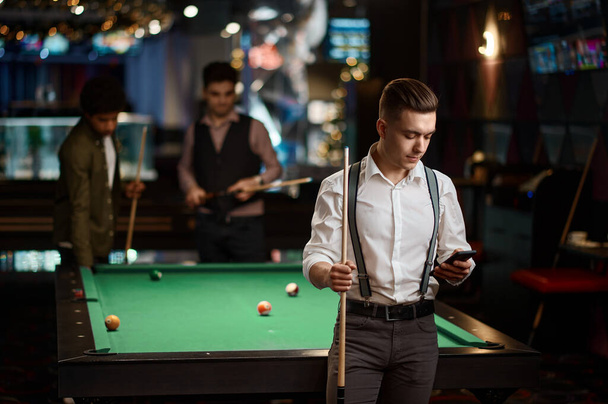 Друзі в барі біля басейну, молодий хлопець відповідає на телефонний дзвінок, а інший чоловік грає в більярд
 - Фото, зображення