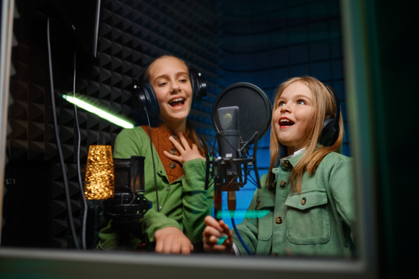 Χαμογελώντας χαρούμενα παιδιά φορώντας ακουστικά τραγουδώντας στο στούντιο ηχογράφησης. Προβολή μέσα από το παράθυρο - Φωτογραφία, εικόνα