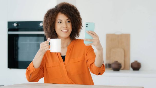 African millennial woman with curly hair girl in orange blouse female vlogger in kitchen at home taking photo on mobile phone holding cup of tea coffee posing recording live stream video on smartphone - Foto, Imagem