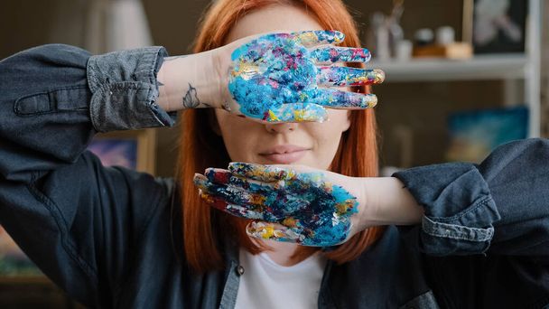 zbliżenie hipster student sztuka klasa pani kaukaski rude włosy dziewczyna artysta kobieta malarz projektant pokazując brudne dłonie w farby trzymając ręce pokrywa twarz z ramionami po rysunek patrząc w aparat wewnątrz - Zdjęcie, obraz