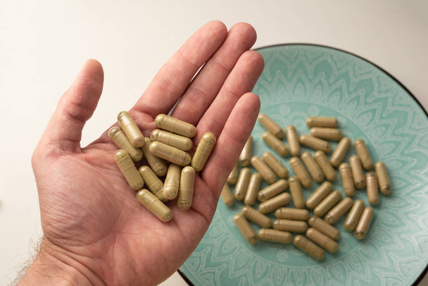 one hand holds some kratom pills with green plate with other kratom pills on background, selective focus - Photo, Image