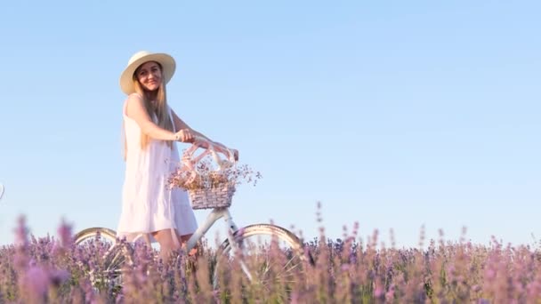 Mulher francesa bonita em um vestido branco e um chapéu branco em um campo de lavanda. Uma loira com uma bicicleta fixa o cabelo. Contra o fundo do céu azul - Filmagem, Vídeo