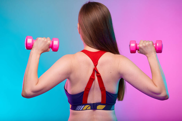 Sporty woman with long hair in fitness sportswear doing exercise with pink dumbbells back view. Female fitness portrait isolated on neon multicolor background. - Fotografie, Obrázek