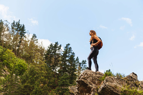ハイキング中に森林や山の風景を考えながら岩の上に立ってリュックを持つ白人ハイカーの女性。週末の野外活動 - 写真・画像