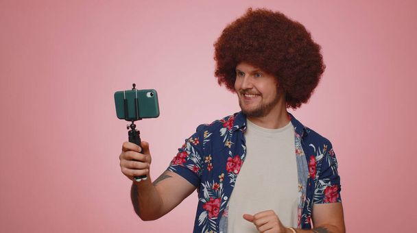 Tourist man in shirt traveler blogger taking selfie on mobile phone selfie stick communicating video call online with subscribers. Young adult guy boy 30s isolated alone on pink studio wall background - 写真・画像