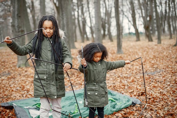 Beautiful little black girls in tent camping in the forest. Two little sisters are puting up a tent in autumn forest. Black girls wearing khaki coats. - 写真・画像