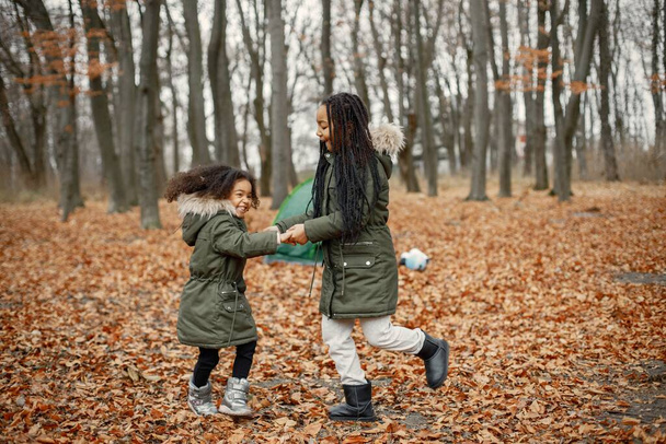 Beautiful little black girls standing near tent in the forest. Two little sisters dancing in autumn forest. Black girls wearing khaki coats. - Foto, afbeelding