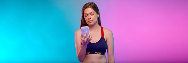 Sporty woman in sportswear using smartphone. Female fitness portrait isolated on neon multicolor background. - Фото, изображение
