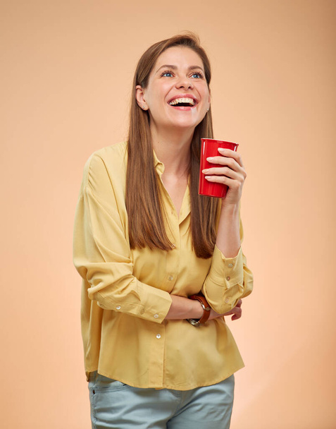Smiling woman looking up, isolated portrait on yellow background. Young lady with long hair holding red coffee cup. - Photo, image
