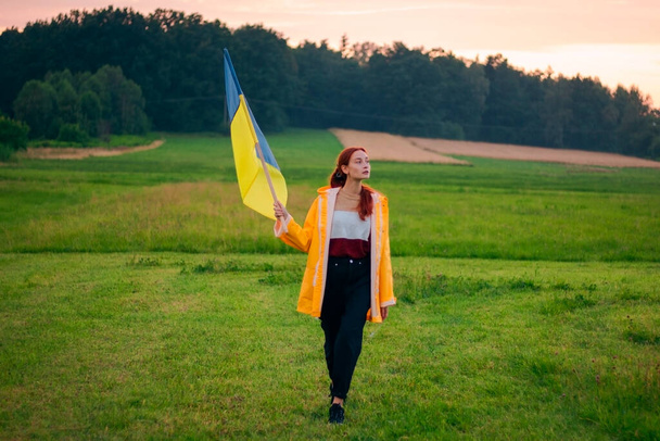 Mujer ucraniana en el campo con una gran bandera de Ucrania. Fotos patrióticas. Heroísmo y libertad. Concepto. Hermosa mujer con un impermeable amarillo. Viajes - Foto, imagen