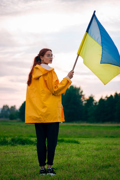 Donna ucraina sul campo con una grande bandiera dell'Ucraina. Foto patriottiche. Eroismo e libertà. Concetto. Bella donna con un impermeabile giallo. Viaggio - Foto, immagini