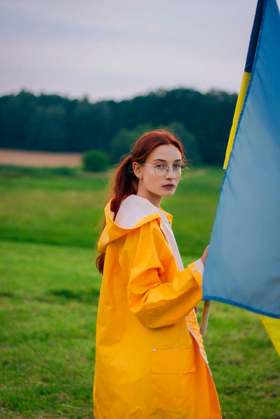 Ukrainische Frau auf dem Feld mit einer großen Fahne der Ukraine. Patriotische Fotos. Heldentum und Freiheit. Konzept. Schöne Frau in einem gelben Regenmantel. Reise - Foto, Bild