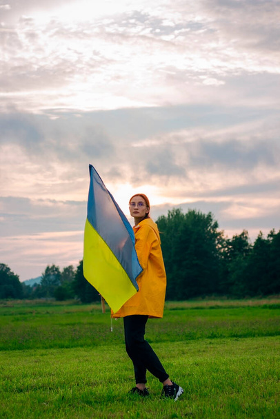 Mujer ucraniana en el campo con una gran bandera de Ucrania. Fotos patrióticas. Heroísmo y libertad. Concepto. Hermosa mujer con un impermeable amarillo. Viajes - Foto, imagen