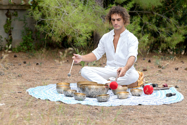 A man playing on a tibetian singing bowls sitting outdoors for meditation. Buddhist healing practices. Sound therapy, alternative medicine. Clearing the space of negative energy. Selective focus - Photo, Image
