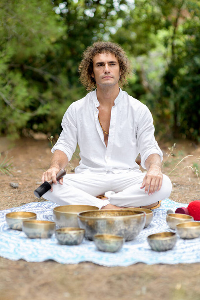 Tibetan singing bowls master sitting outdoors with many tibetan singing bowls, getting ready for meditation. A man wearing in white practice sound therapy, Buddhist healing practices. Selective focus - Photo, Image