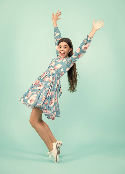 Excited teenager, amazed and cheerful emotions. Happiness, freedom, motion and child. Young teenager girl wear summer dress jumping in air over blue background - 写真・画像