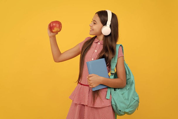 Gülümseyen genç kız sırt çantası taşıyor. Okula dönüyorum. Bilgi günü. Eğitim konsepti. Öğlen yemeği için kulaklıklı, çalışma kitabı ve elmalı bir çocuk. 1 Eylül, sağlıklı bir çocukluk. Okul çantalı çocuk. müzik - Fotoğraf, Görsel