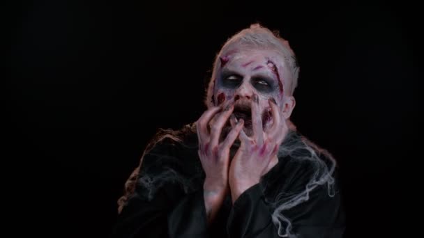 Zombie-Mann mit Make-up mit Wundnarben und weißen Kontaktlinsen, der krampfhaft in die Kamera blickt und versucht, im dunklen Studioraum zu erschrecken. Finsteres Totenmonster. Halloween, Dreharbeiten, Bühnenkonzept - Filmmaterial, Video