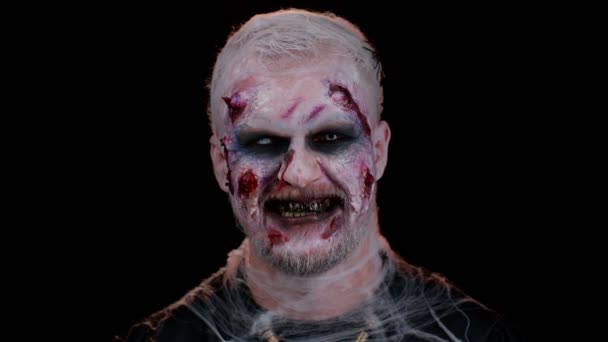 Pahaenteinen mies, jolla on kauhea pelottava Halloween-zombie-meikki puvussa, joka näyttää likaisilta hampailta, katsoen kameraa yrittäen pelotella. Kuollut kaveri haavoittunut verinen arvet kasvot eristetty mustalla pohjalla - Materiaali, video