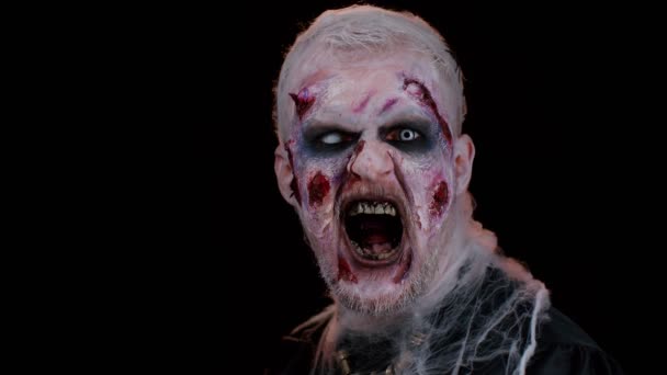 Zombie man with makeup with fake wounds scars and white contact lenses looking at camera and screaming, trying to scare in dark studio room. Sinister dead guy. Halloween, filming, staging concept - Footage, Video