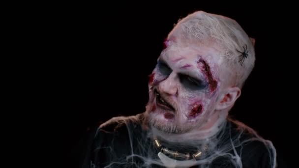 Beängstigende Mann mit Halloween-Zombie blutig verwundet Make-up, versuchen zu erschrecken, Gesichtsausdruck. Krämpfe. Finsterer Untoter isoliert auf schwarzem Hintergrund im Studio. Voodoo-Rituale. Modische Körperkunst - Filmmaterial, Video