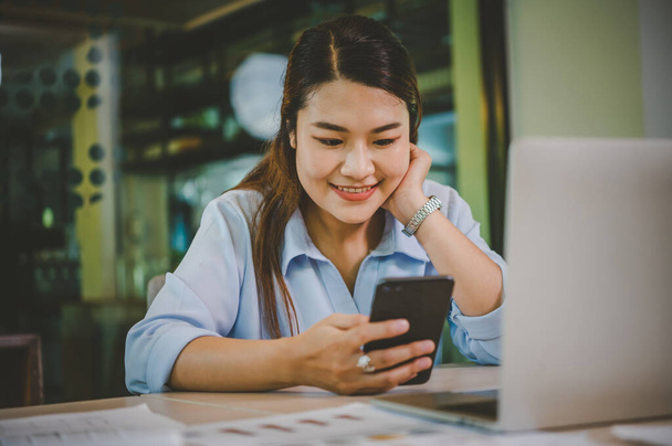 Mujer asiática de negocios sentada en su escritorio con computadora portátil utilizando el teléfono móvil durante la comprobación de un correo electrónico o redes sociales en Internet.  - Foto, imagen
