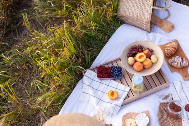 White blanket with fruits and pastries, lemonade, cookies, cheese and straw bag with baguette. Concept of having picnic in a city park during summer holidays or weekends. Copy space. - Foto, imagen