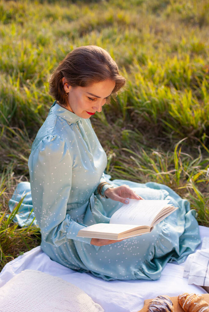 A woman in a long summer dress with short hair sitting on a white blanket with fruits and pastries and reading the book. Concept of having picnic in a city park during summer holidays or weekends.  - Photo, Image