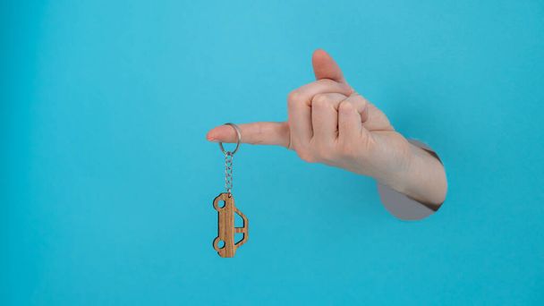 Womans hand holding a wooden keychain in the shape of a car on a blue background - Foto, Imagen