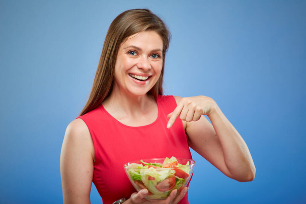 Mladá usměvavá dáma v červených šatech drží salátovou mísu a ukazuje prstem, izolovaný portrét na modrém pozadí. - Fotografie, Obrázek
