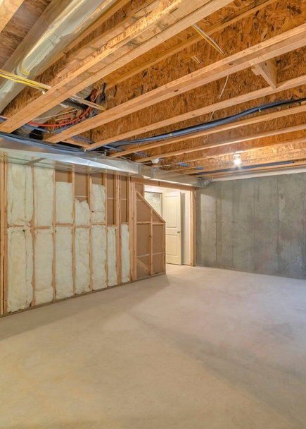 Vertical Unfinished basement with a plastic vapor barrier on the wall. Empty basement interior with wooden frameworks and insulated wall on the left and a window on the right. - Photo, Image