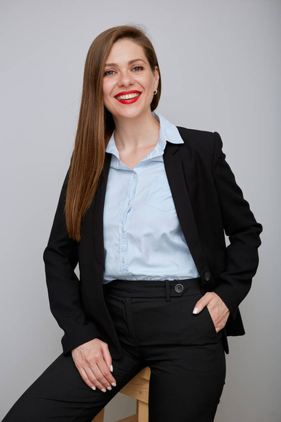 Smiling adult woman in black business suit sitting on tall stool. Young business woman with hand in pocket isolated portrait. - Φωτογραφία, εικόνα
