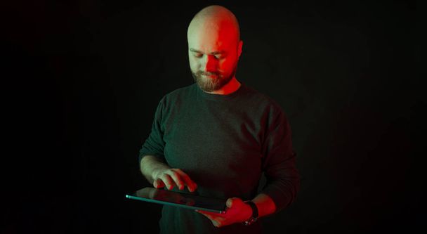 Content balding man works with the tablet over black background with red light. Panorama studio shot. - Photo, Image