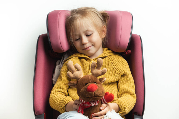 Cute little girl holding stuffed Rudolph Red-Nosed Reindeer toy while sitting in a safety car seat. Kid safety while traveling by car. Isolated on white. Merry Christmas. Holiday adventure concept - Foto, Imagen