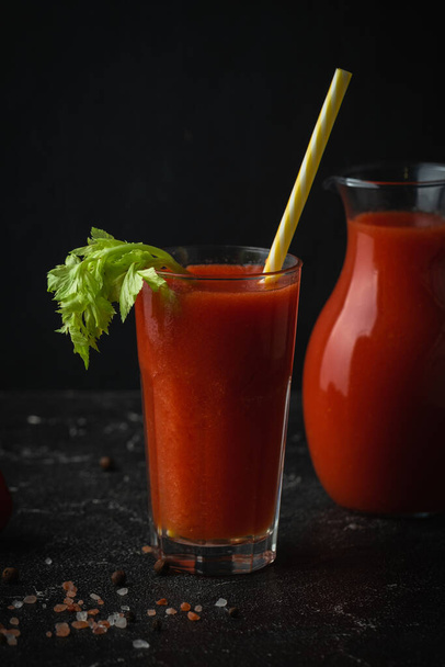 Fresh tomato juice in a glass with a straw on the black background. Red tomato juice cocktail with tomatoes and celery - Fotó, kép