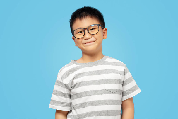Smart kid with smile, wearing eyeglasses and tshirt looking at camera on blue background - Fotó, kép