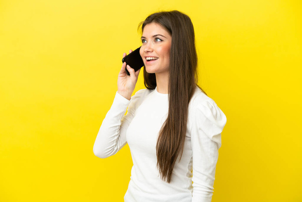Young caucasian woman isolated on yellow background keeping a conversation with the mobile phone - Фото, изображение