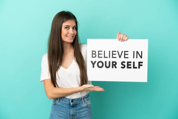 Young caucasian woman isolated on blue background holding a placard with text Believe In Your Self and pointing it - 写真・画像
