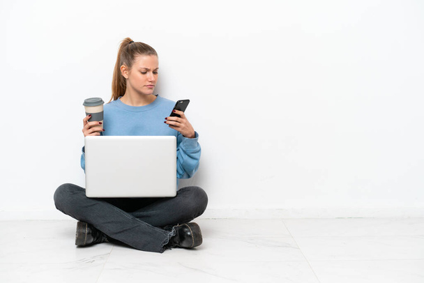 Молода жінка з ноутбуком, що сидить на підлозі, тримає каву, щоб забрати і мобільний
 - Фото, зображення