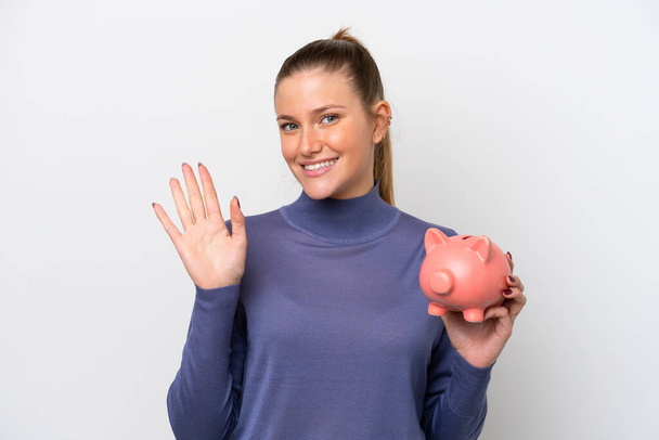 Young caucasian woman holding a piggybank isolated on white background saluting with hand with happy expression - Photo, Image