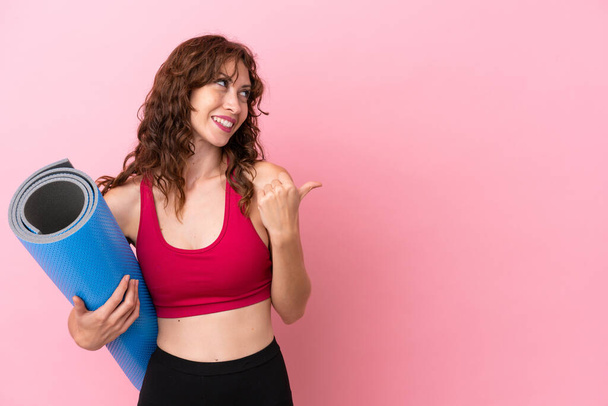 Jeune femme sportive allant à des cours de yoga tout en tenant un tapis isolé sur fond rose pointant vers le côté pour présenter un produit - Photo, image