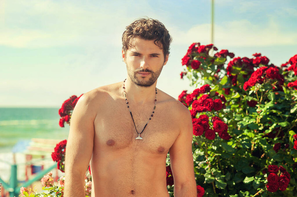 Handsome naked guy standing near rose bush on sea resort - Photo, Image
