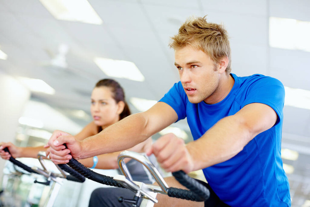 Man on exercise bike. Young man cycling at the gym with woman in background - Fotoğraf, Görsel