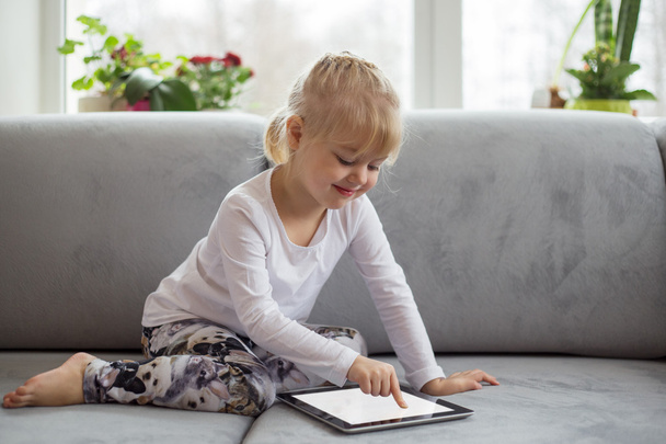 Умная маленькая девочка с помощью планшетного компьютера, сидя на диване в гостиной на дому - Фото, изображение