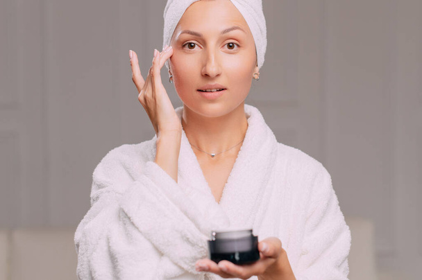 Young attractive woman in a bathrobe applies cream on her face in the bathroom. - Φωτογραφία, εικόνα